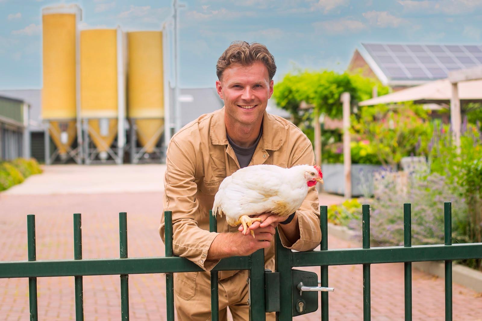 De boer bij het hek van het erf, met een kip op de arm en de silo's op de achtergrond